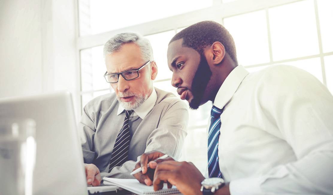 Concept for teamwork and knowledge transfer Photo of aged businessman and his colleague. Businessmen working in office with big window. Men using laptop